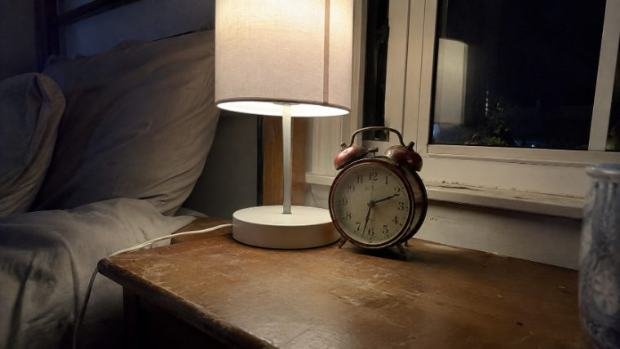 A bedside lamp and an old alarm clock on a nightstand at night.